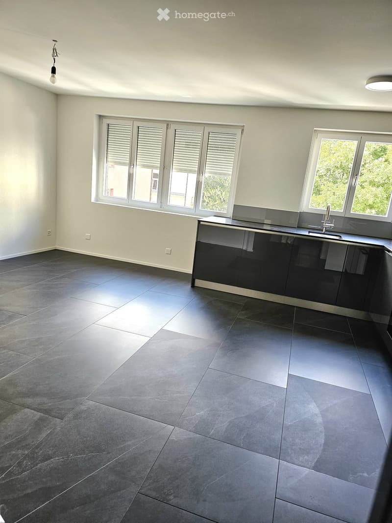 Empty kitchen with dark grey tiled floor, white walls, and large windows with blinds. A modern kitchen counter with a sink and faucet is visible, but no other furniture or appliances are present.
