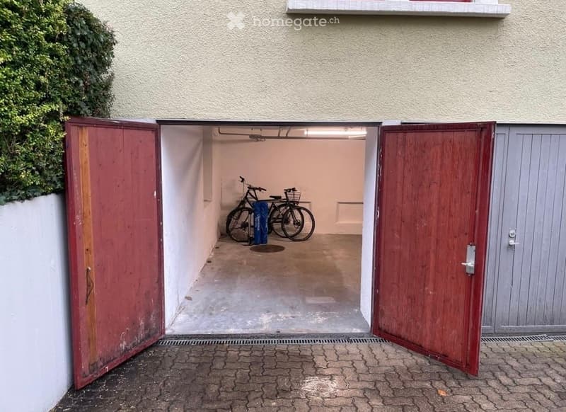 Garage with two bicycles inside, red wooden doors, concrete floor, and a paved entrance area.
