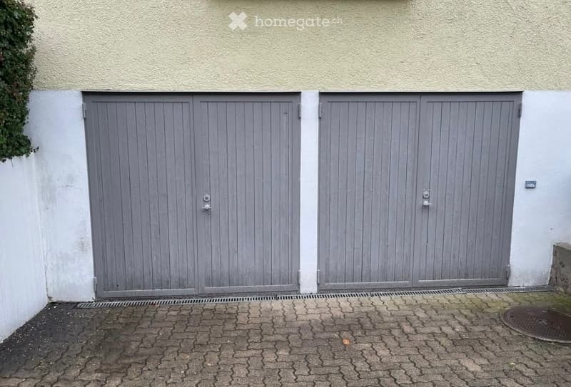 Two grey garage doors on a beige building exterior, with a paved brick driveway and a small green plant on the left side.