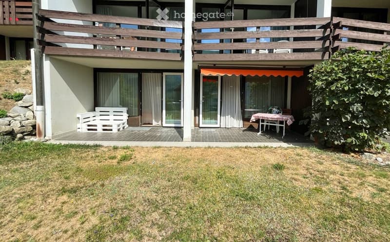 Outdoor patio area with a wooden bench, a small table with chairs, and an orange awning. The patio is connected to a house with large windows and a wooden balcony above.