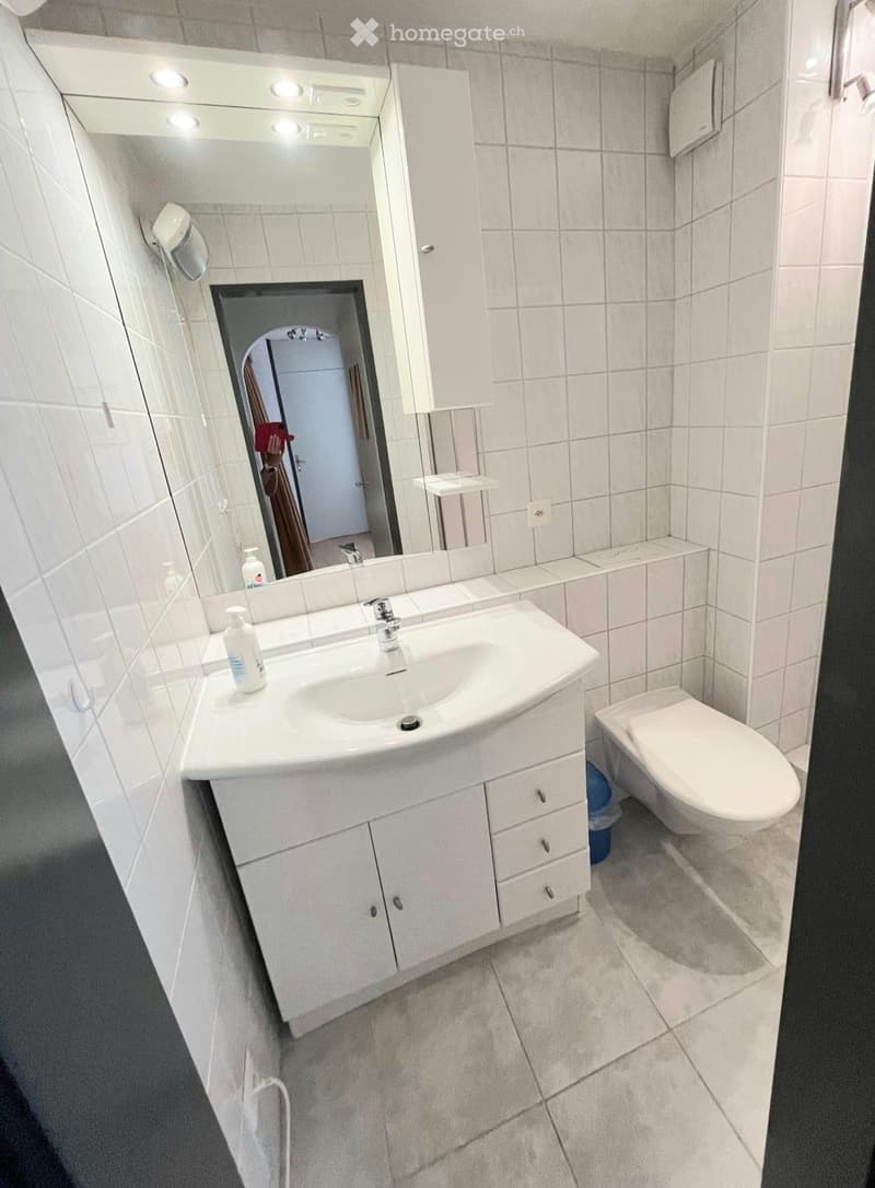 Modern bathroom with white tiled walls and floor, a white sink with a cabinet below, a toilet, and a large mirror with built-in lighting above the sink.