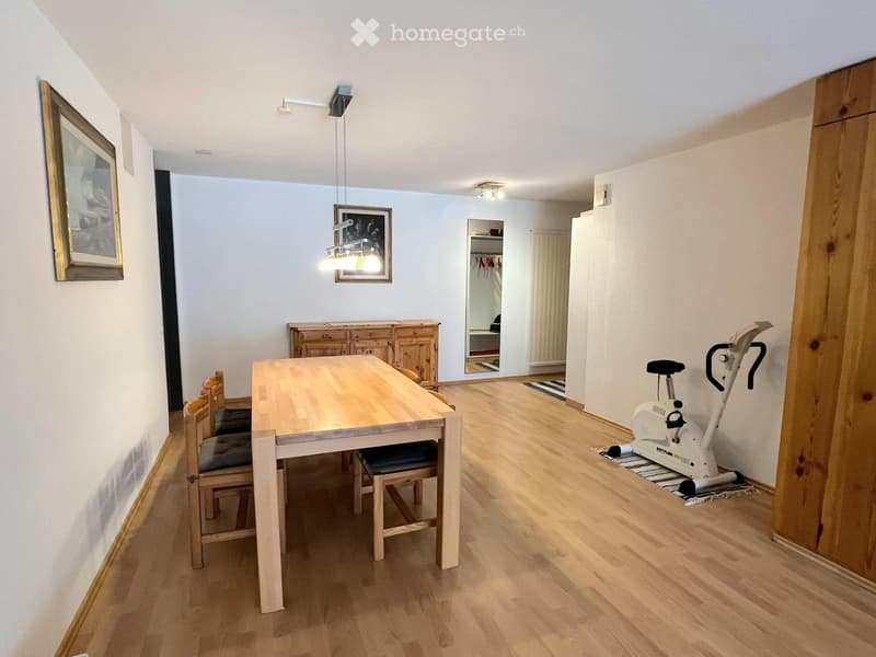 Bright, open room with wooden flooring, a dining table with chairs, a wooden cabinet, an exercise bike, and a mirror in the background.
