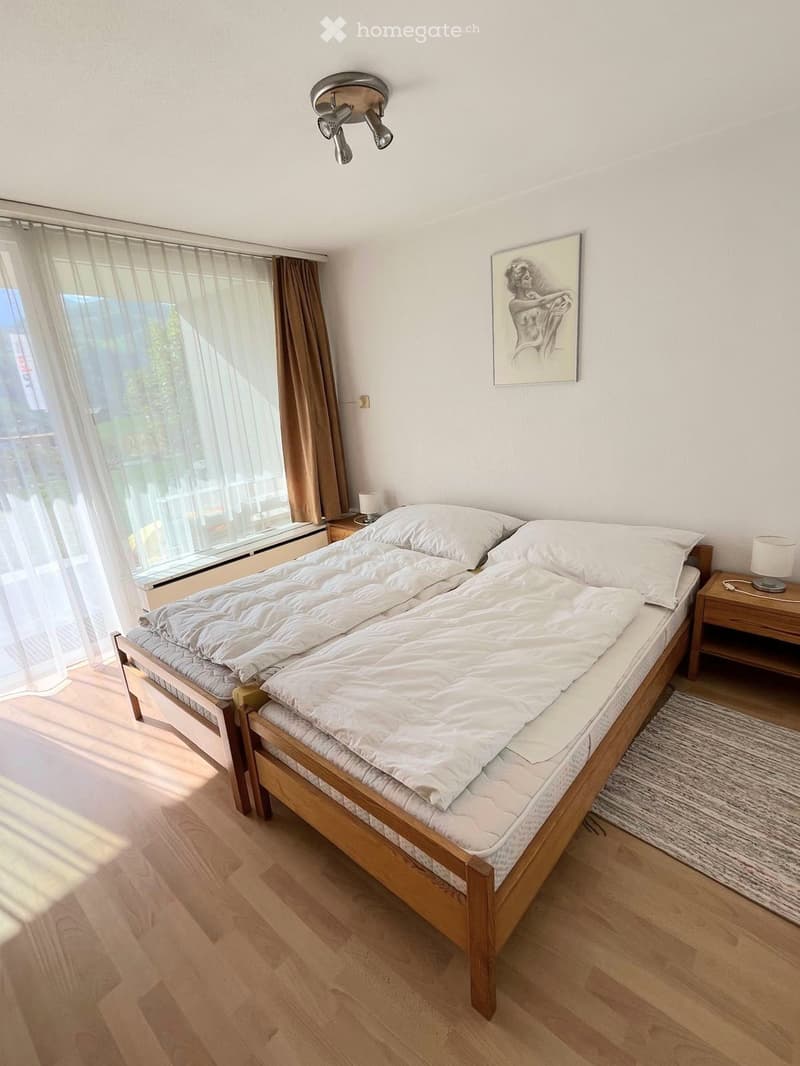 Cozy bedroom with a wooden bed, white bedding, nightstand, lamp, and a framed picture on the wall. Large window with sheer curtains and a view of the outside.