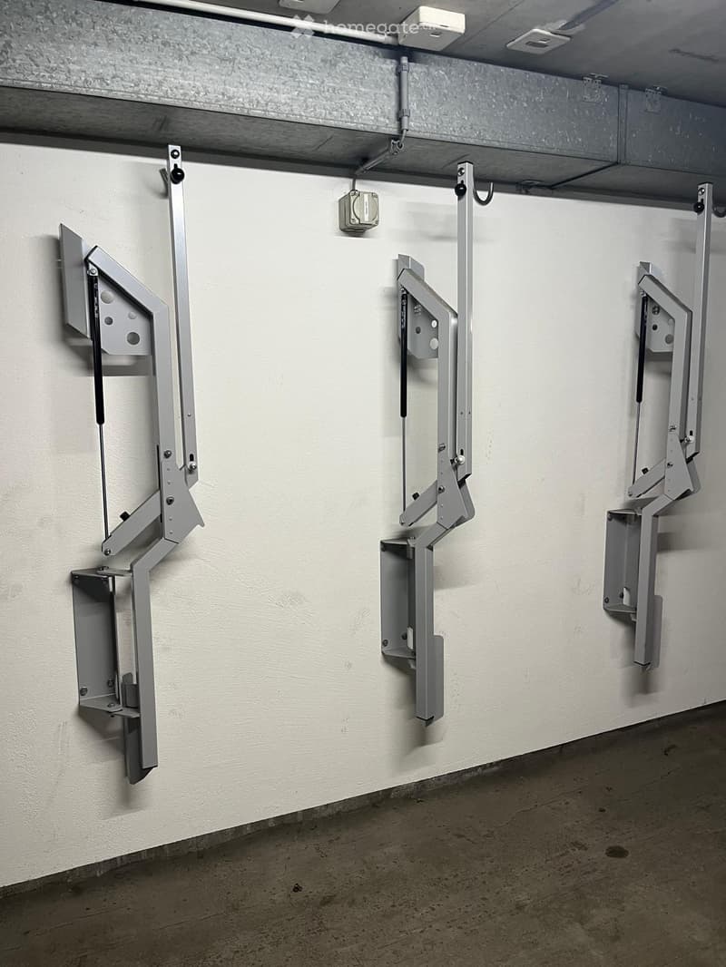 A concrete-floored hallway with three wall-mounted metal brackets or holders, each featuring a rectangular, open-fronted compartment with visible screws and bolts.