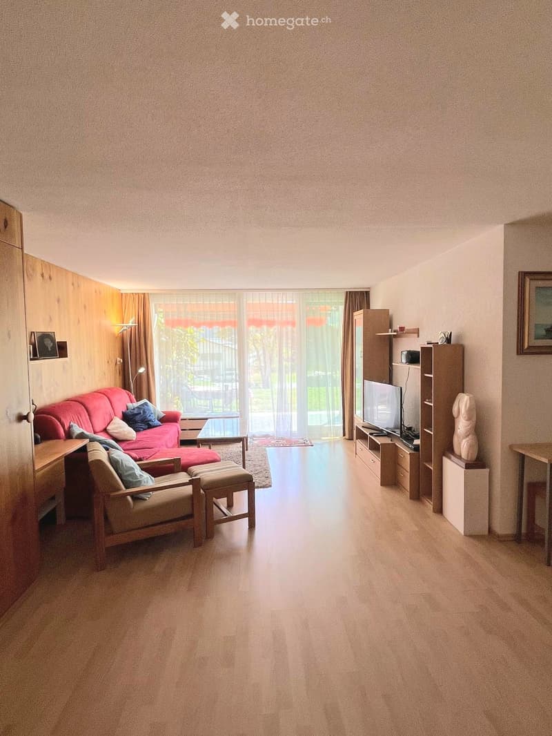 Bright living room with a red sofa, wooden chair, ottoman, coffee table, TV, shelves, and a statue. Large windows with curtains provide natural light.