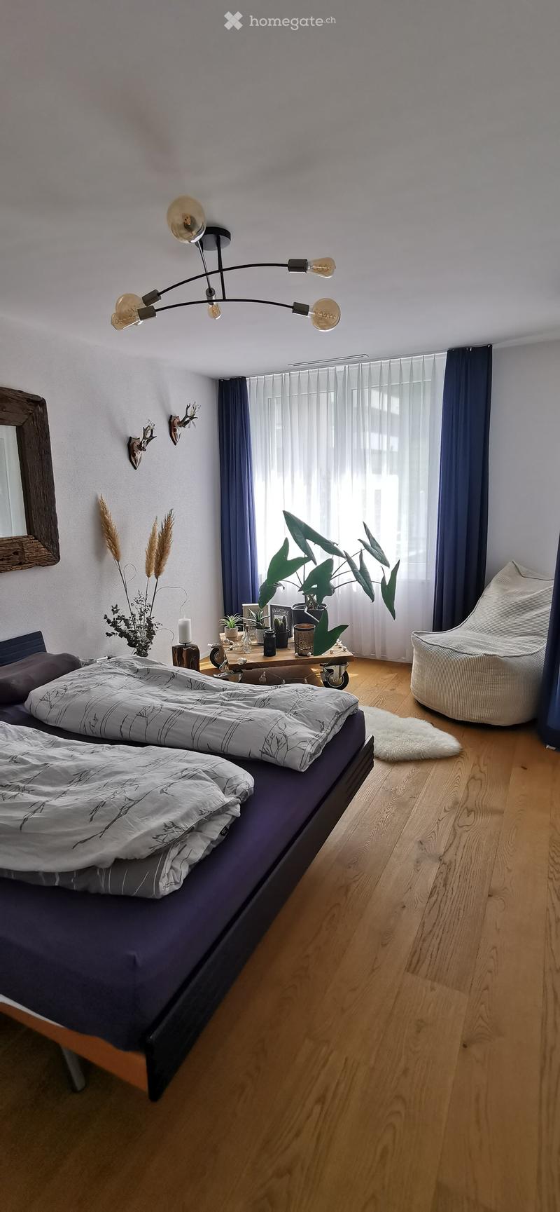 Modern bedroom with a double bed, nightstand, side table, large window with curtains, and decorative plants.