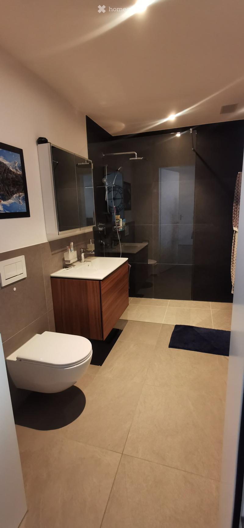 Modern bathroom with beige tiles, a white toilet, a wooden vanity with a sink, a mirror, and a glass-enclosed shower area.