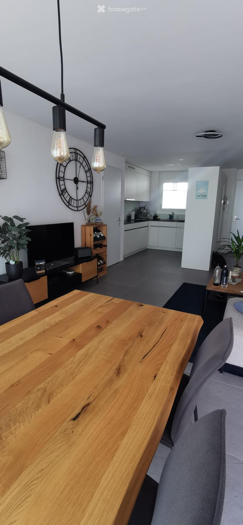 A modern living and dining area with a large wooden dining table, chairs, a TV, a small kitchen area, and decorative elements like a clock and plants.