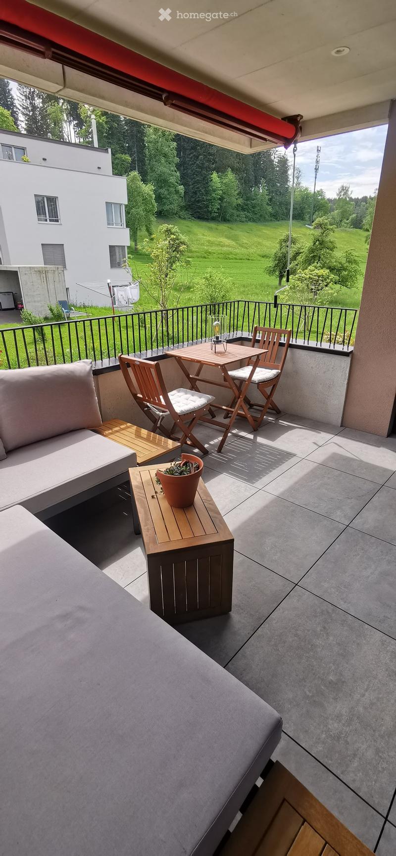 Balcony with a grey sofa, wooden table, chairs, potted plant, and a bench, overlooking a green lawn and trees.