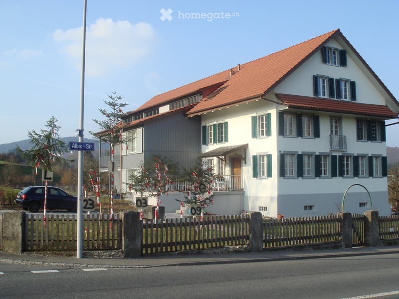 Two-story house with red-tiled roof, white walls, green shutters, and a small balcony. The house is surrounded by a wooden fence and trees.