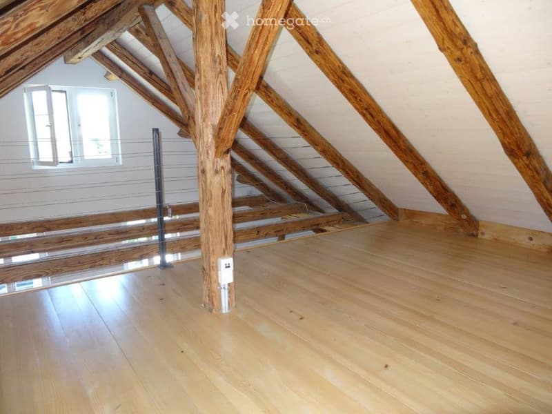 Empty attic space with wooden beams, exposed wooden ceiling, and wooden floor. A window on one side provides natural light.