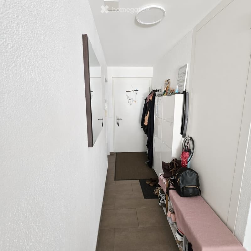 A clean, modern hallway with white walls, a tiled floor, and a small bench with shoes and bags. A coat rack with jackets and a mirror are visible.