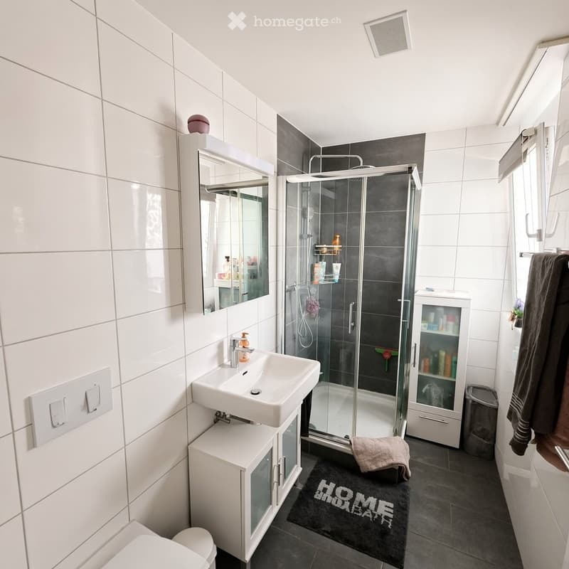 Modern bathroom with white tiles, a white sink with a cabinet, a large mirror, a glass-enclosed shower, a toilet, and a towel on the floor.