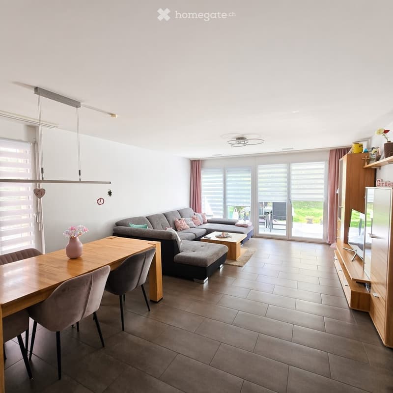 Bright living room with a grey sectional sofa, coffee table, ottoman, dining table with chairs, and a TV stand. Large windows and sliding glass doors lead to a balcony.
