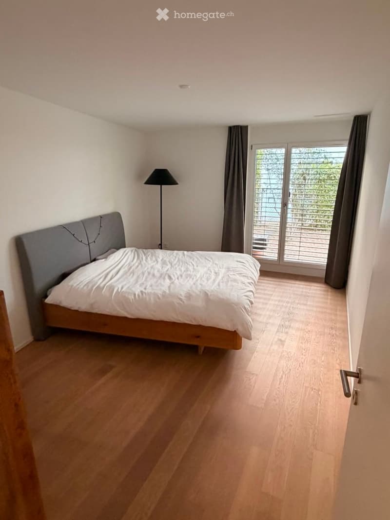 Modern bedroom with light wooden flooring, a grey upholstered bed, and a tall floor lamp near the window. The room has large windows with dark curtains and a door leading to a balcony.