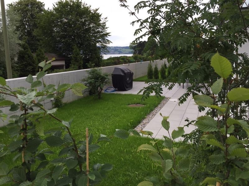 Well-maintained garden with a lawn, gravel pathway, and a barbecue cover, surrounded by trees and a view of a body of water.