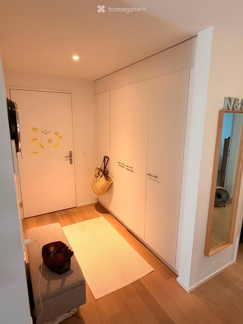 A small, modern hallway with a wooden floor, white walls, built-in wardrobes, a small stool with a handbag, a woven basket hanging on the wall, and a mirror with a fan visible in the reflection.