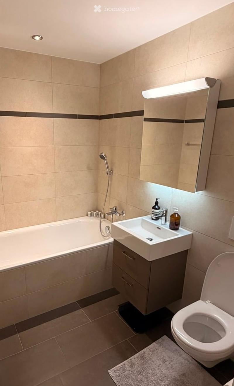 Modern bathroom with beige tiled walls, a bathtub, a sink with a cabinet, a toilet, a mirror with built-in lighting, and a shower hose.