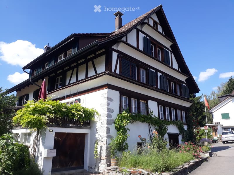 Traditional two-story house with white and brown timber framing, multiple windows, and a stone base. Features include a garage entrance, balcony with greenery, and a well-maintained garden.