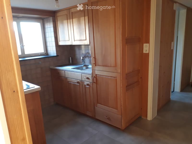 The kitchen features wooden cabinets, a sink with a faucet, and tiled walls. The space is well-lit with a window providing natural light.