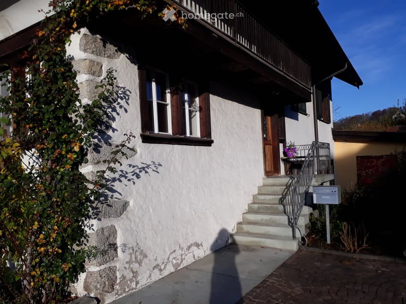 Two-story house with white plaster walls, wooden shutters, stone accents, and a small staircase leading to the entrance.