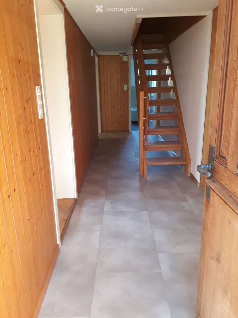 A narrow hallway with wooden paneling on one side, a staircase leading upwards, and tiled flooring.