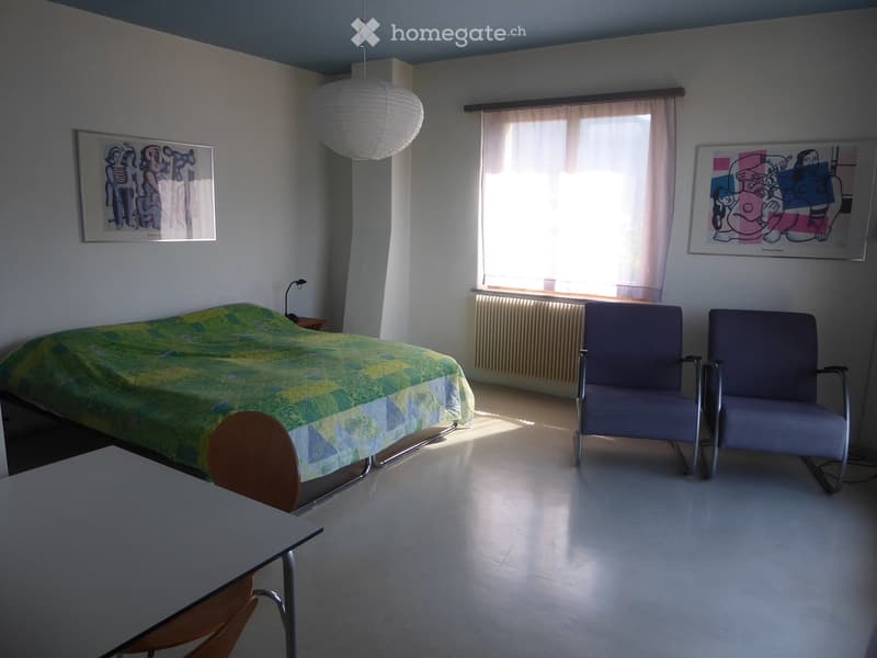 Bright bedroom with a single bed, two chairs, a small table, and artwork on the walls. Natural light streams in from a window with curtains.