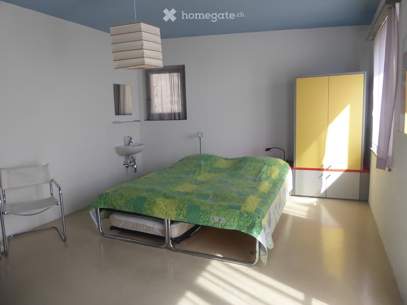 Bright bedroom with a single bed, a chair, a sink, a mirror, and a wardrobe. The room has white walls and a light-colored floor.