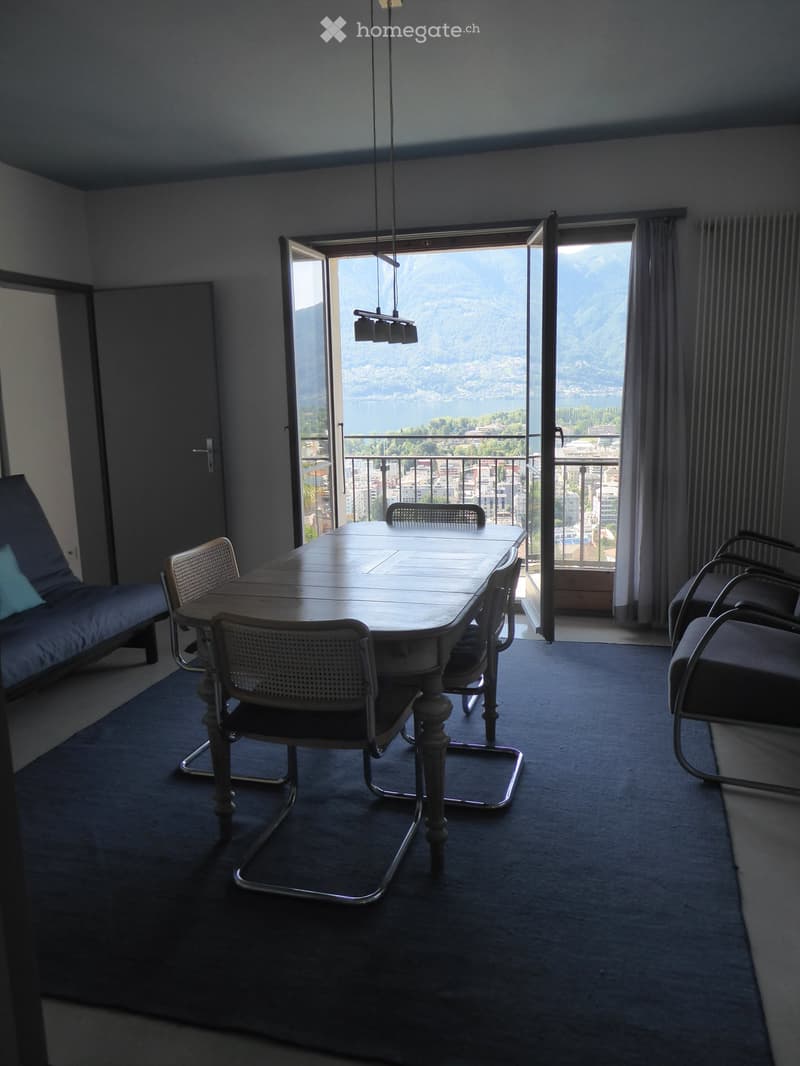 Furnished room with a sofa, dining table, and chairs, large windows offering a scenic view of a lake and mountains.