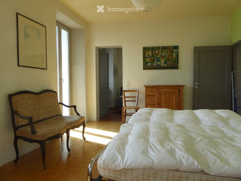 Bright bedroom with wooden flooring, a metal-framed bed with white bedding, a patterned sofa, a wooden chair, and a wooden cabinet with drawers.