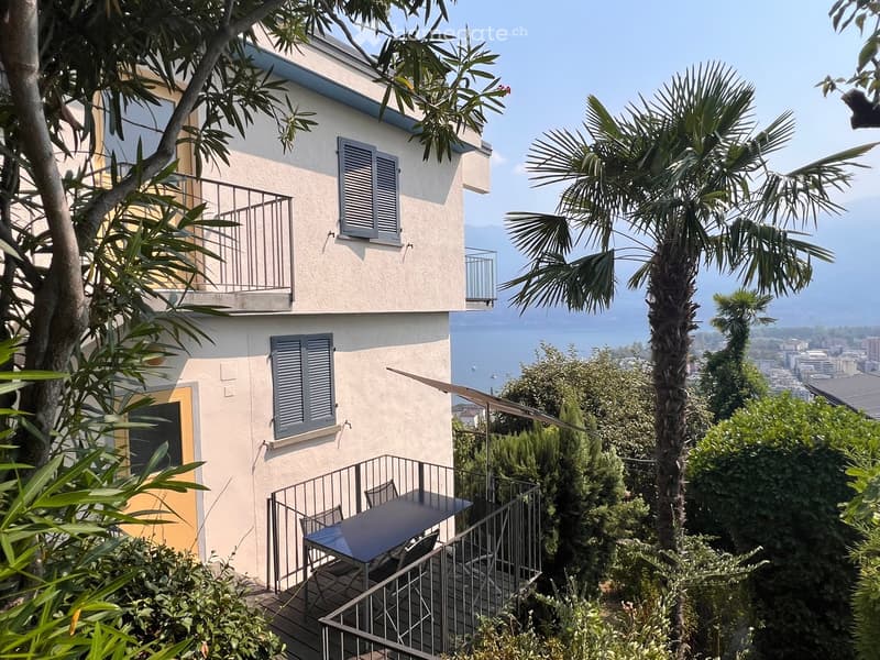 Two-story building with balconies, surrounded by lush greenery and trees, offering a scenic view of the water and distant cityscape.