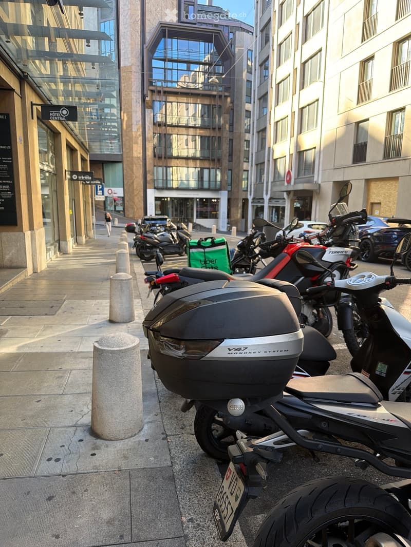 A row of parked motorcycles and scooters on a city sidewalk, with modern buildings and a clear sky in the background.