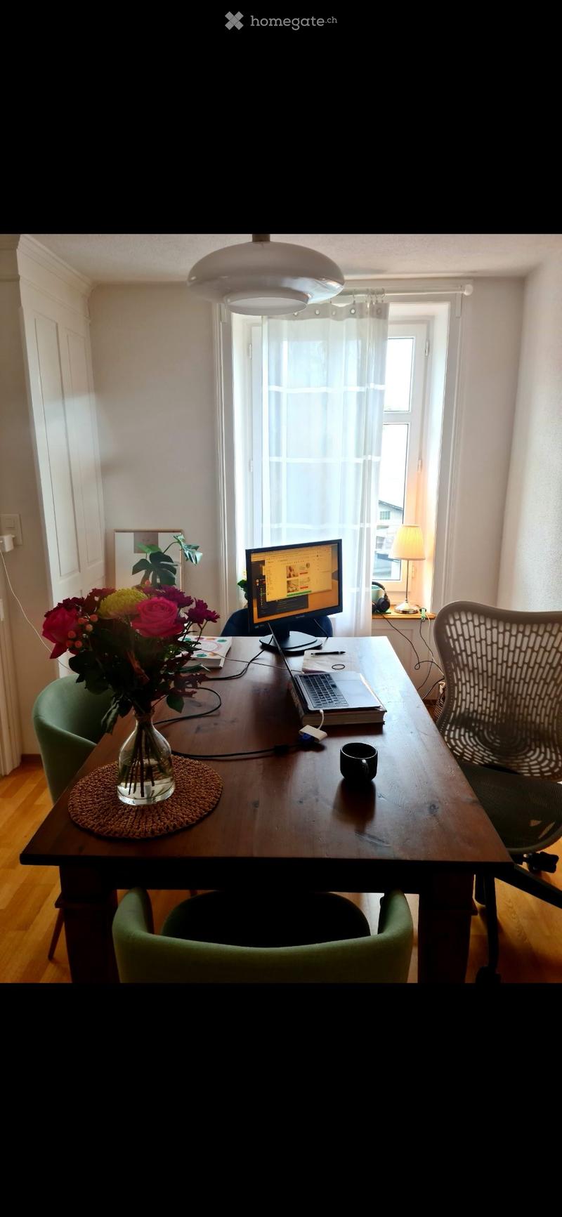 A well-lit room with a wooden table, green chairs, a laptop, a vase with flowers, and a lamp on a side table.