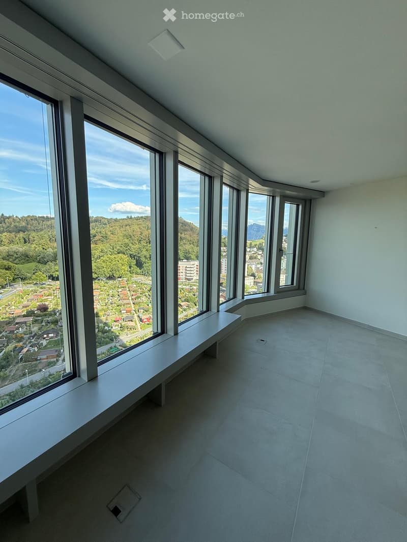 Empty room with large windows offering a scenic view of a town and surrounding greenery. The room features a smooth tiled floor and a built-in bench along one wall.