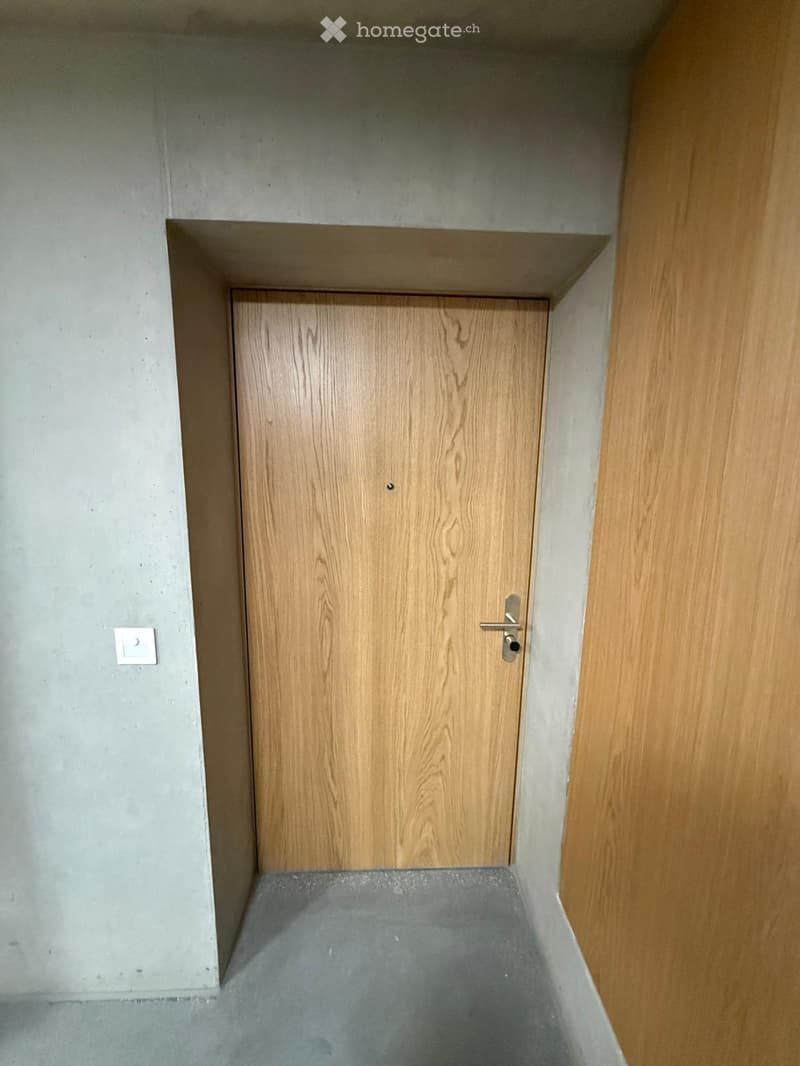 A narrow hallway with a wooden door at the end, concrete walls, and a concrete floor.