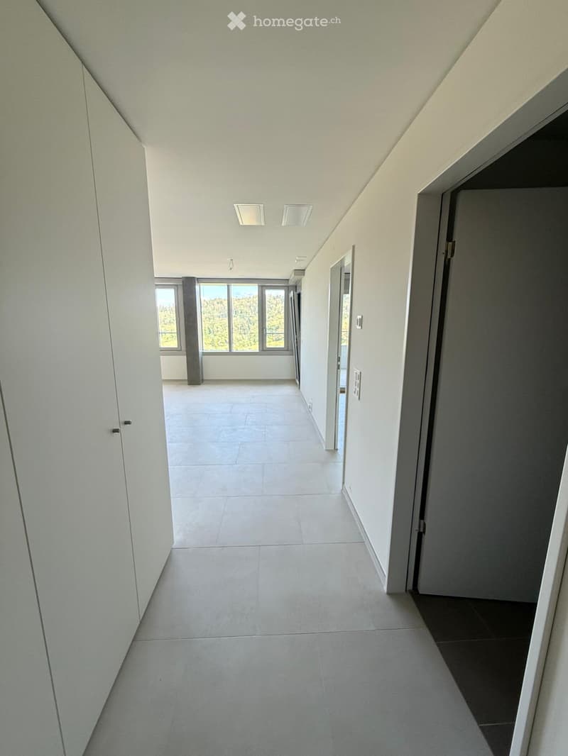 Empty hallway with white walls, tiled floor, and recessed ceiling lights. Features a large window at the end and several doors.