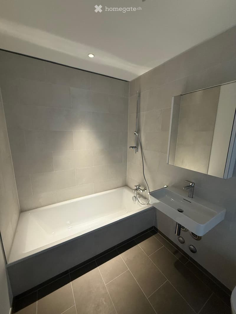 Modern bathroom with a white bathtub, a sink with a modern faucet, a glass-enclosed shower area, and a mirror above the sink. The walls and floor are tiled in neutral tones.