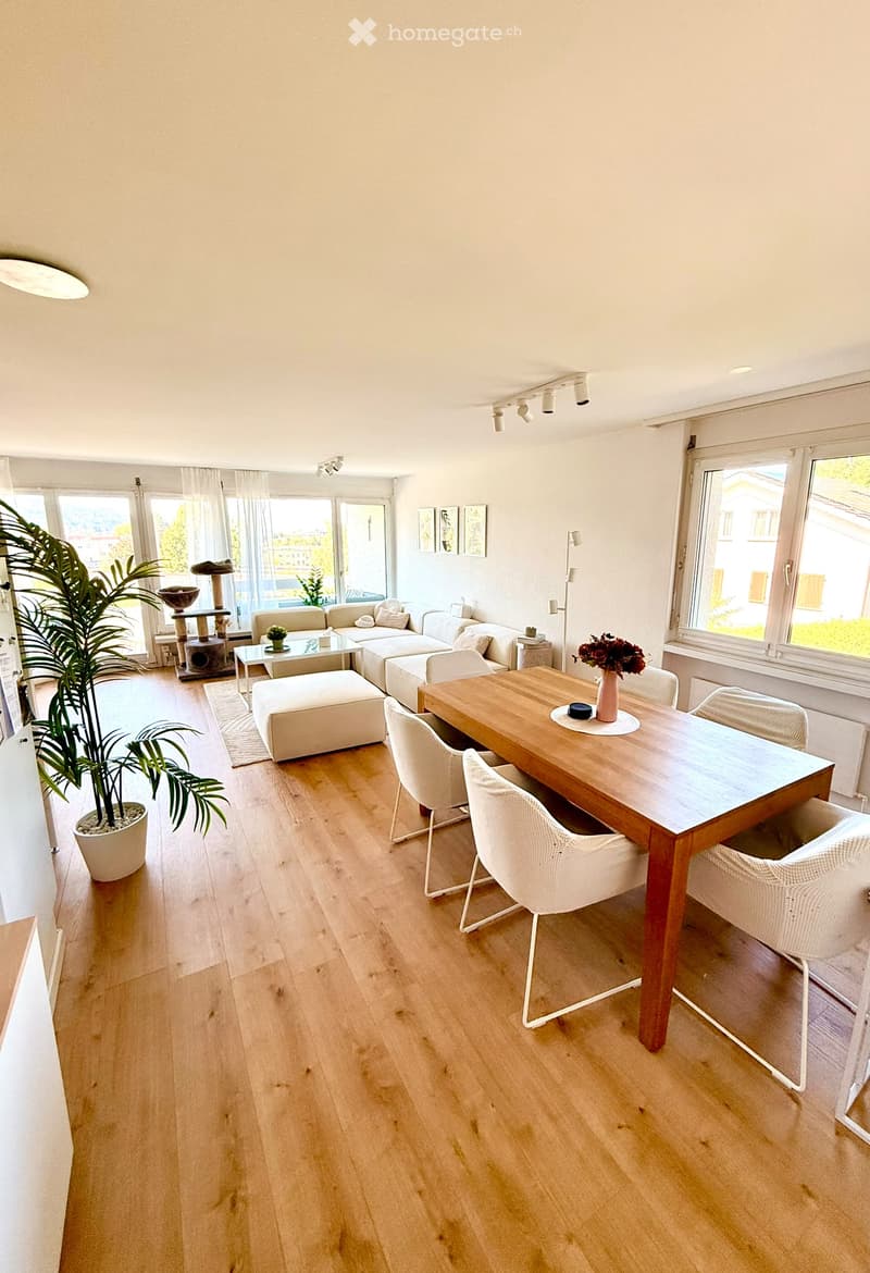 Bright living and dining area with light wooden flooring, large windows, a white sofa, ottoman, coffee table, dining table with chairs, cat tree, and decorative plant.