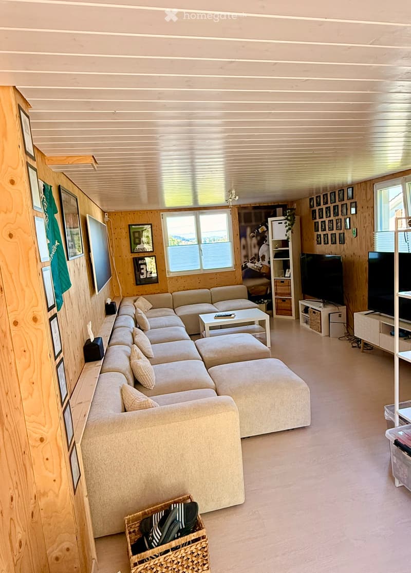 Cozy living room with a large sectional sofa, coffee table, ottoman, TV stand, and a woven basket on the floor. The room features wooden walls and large windows.