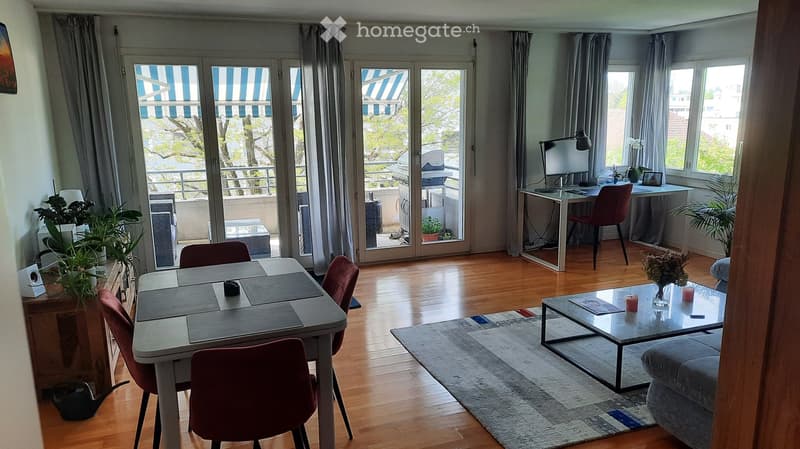 Open-plan living and dining area with wooden flooring, large windows, and a balcony. Features a sofa, coffee table, dining table with chairs, desk with a computer, and decorative plants.