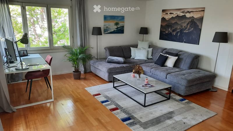 Modern living room with grey sectional sofa, coffee table, glass desk, chair, floor lamps, and artwork on the walls.