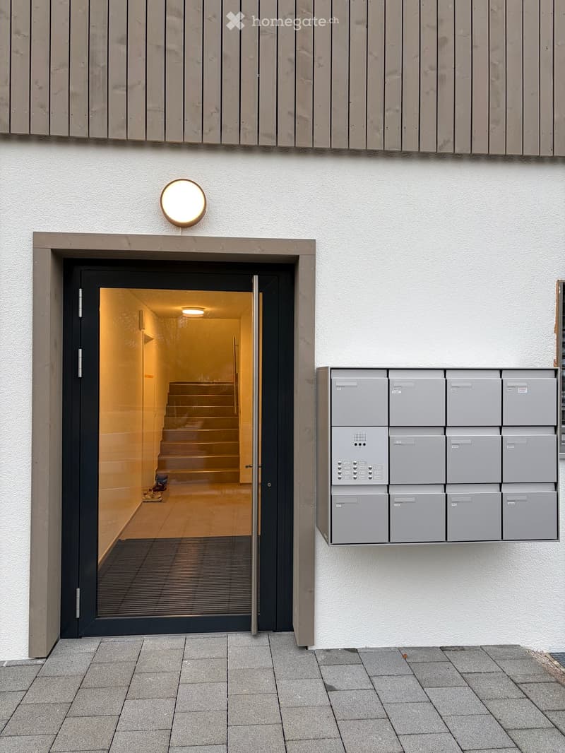 Entrance to a building with a modern design, featuring a glass door, a staircase inside, and mailboxes on the wall.