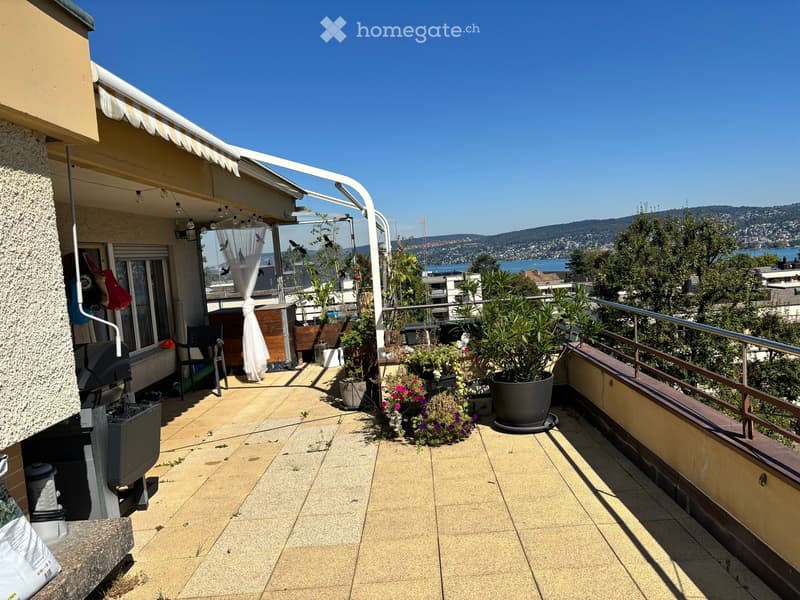 Balcony with tiled floor, metal railing, potted plants, and a view of the sea and distant hills.