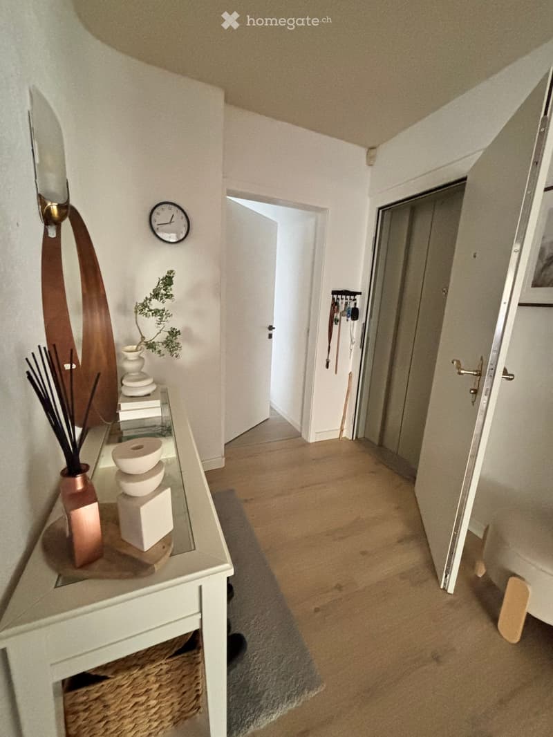 A well-lit hallway with wooden flooring, a small console table with decorative items, a clock on the wall, and an open doorway leading to another room.