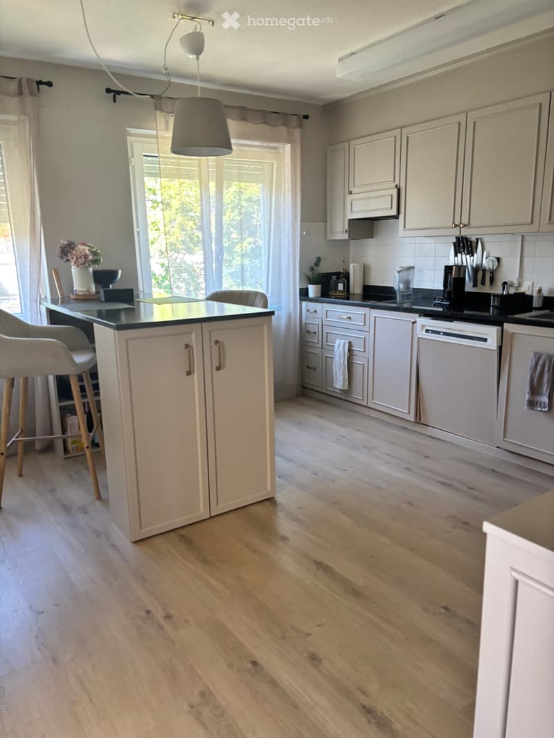 Modern kitchen with light-colored cabinets, wooden flooring, and a central island with a black countertop. Appliances include a dishwasher, coffee maker, and blender. A vase with flowers sits on the counter, and a hanging pendant light illuminates the spa