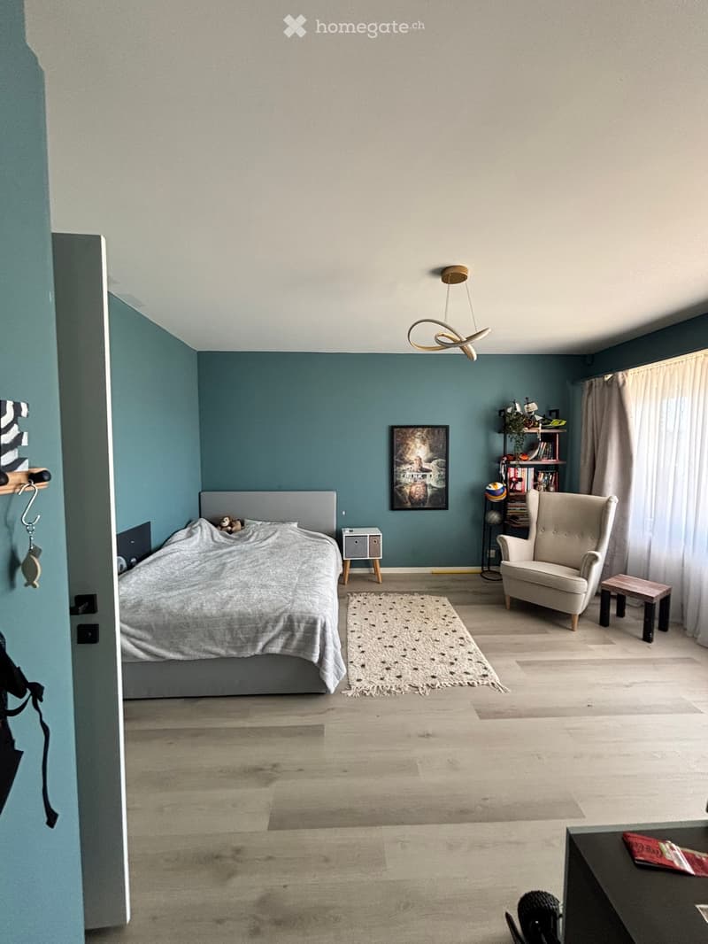 Modern bedroom with a grey bed, a beige armchair, a small wooden table, a shelf with books and decorative items, a framed picture, and a patterned rug on a light wooden floor.