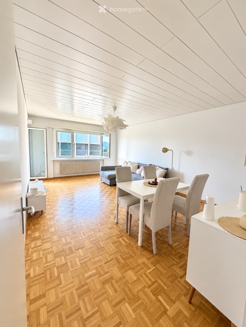 Bright living and dining area with wooden herringbone flooring, white dining table with four chairs, grey sofa with throw pillows, side table, and modern lighting.