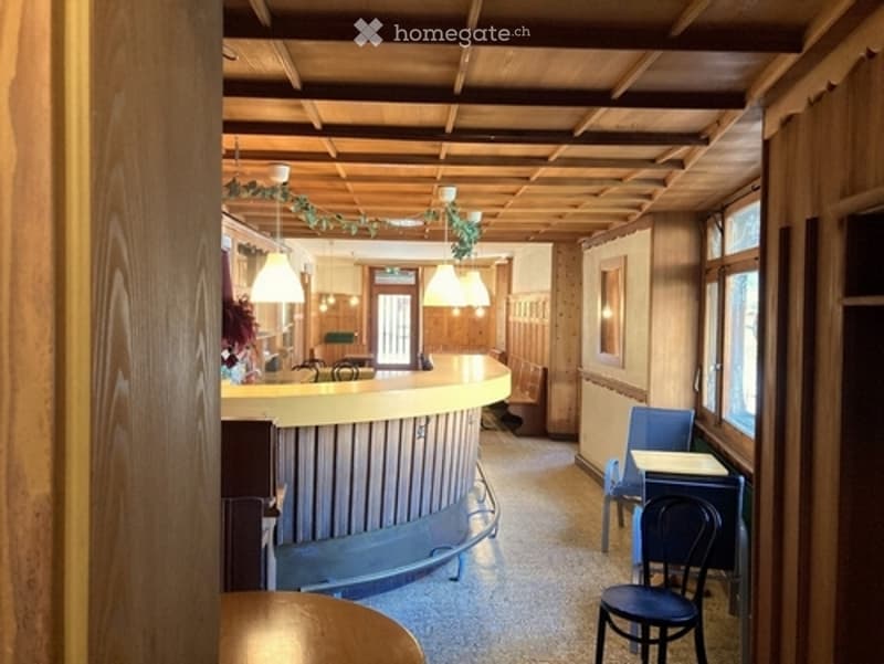 A reception area with a curved wooden desk, chairs, and pendant lights. The space features wooden paneling on the ceiling and walls, with a small plant on the desk and a window letting in natural light.