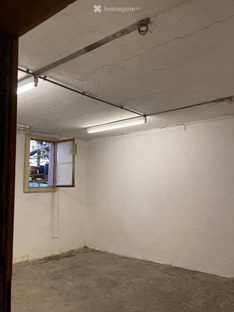 Empty room with bare white walls, concrete floor, and a single window with wooden frame. Exposed pipes and electrical wiring are visible on the ceiling.