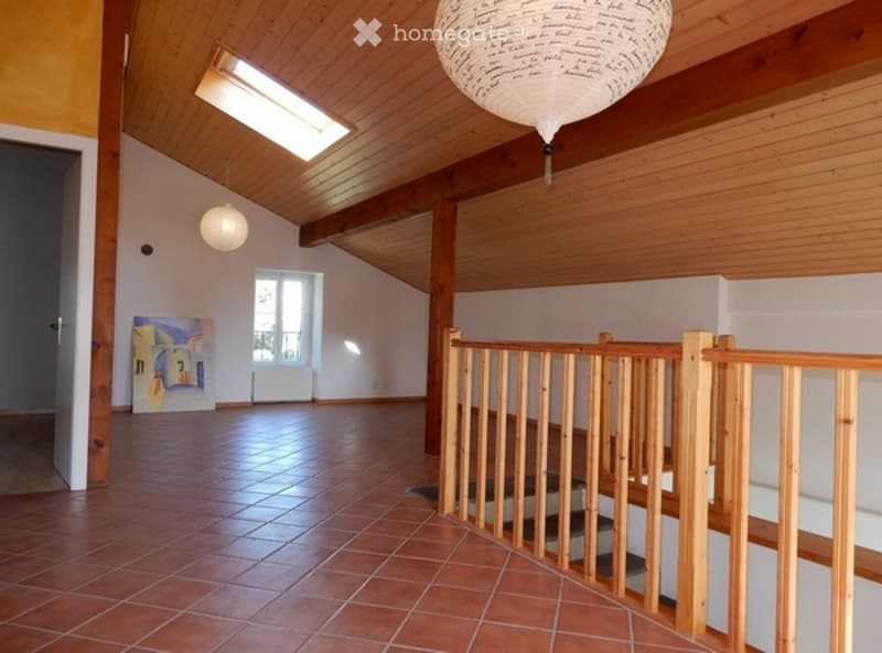 Bright, open living space with wooden ceiling beams, exposed wooden beams, and a staircase leading upwards. The room features a skylight, a large window, and a modern pendant light. The floor is covered with terracotta tiles.